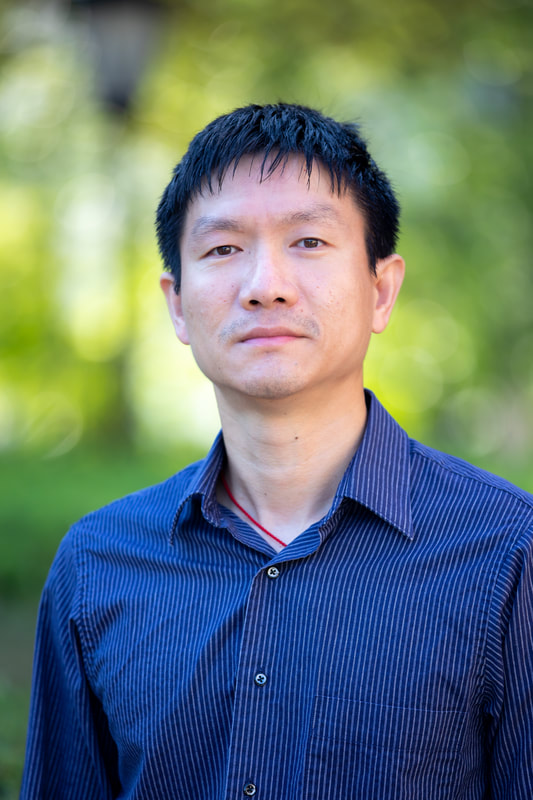 Professional headshot of Dr. Junfei Lu in front of blurred outdoor background with trees and sunlight